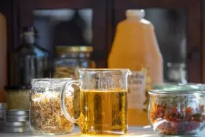 Close-up of jojoba oil in a glass beaker, with dried herbs and flowers in the background.