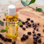 bottle of hobacare jojoba in bottle on wooden board with jojoba seeds sprinkled around and jojoba branch in upper right corner