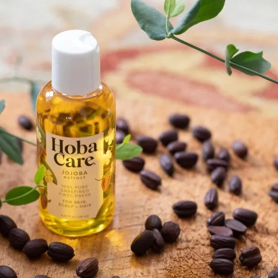 bottle of hobacare jojoba in bottle on wooden board with jojoba seeds sprinkled around and jojoba branch in upper right corner