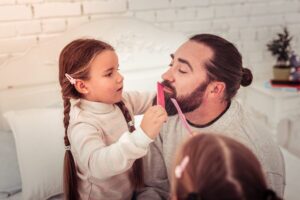 little girl with brown braids combing dad's brown beard with a pink comb to promote that jojoba is the best beard oil
