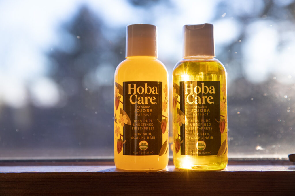two bottles of golden HobaCare jojoba oil in a windowsill with the one on the left cloudy and solid from being exposed to temperatures below 50 degrees, bottle not he right is still in its liquid state and is clear and golden