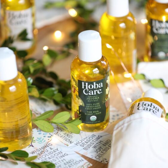 bottles of golden organic HobaCare jojoba surrounded by golden fairy lights, muslin gift bags, and greenery