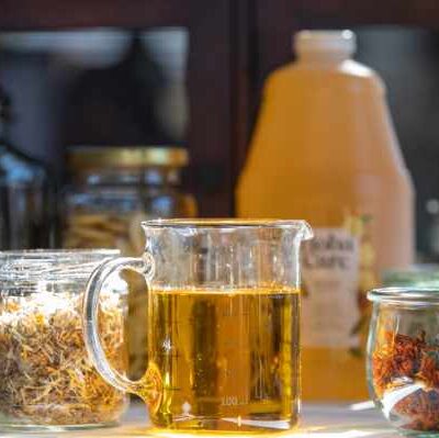 Close-up of jojoba oil in a glass beaker, with dried herbs and flowers in the background.