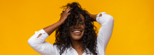 woman in white shirt with hands in textured hair standing in front of a bright yellow background