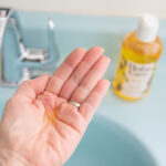 golden jojoba oil in an open palm with a light blue sink in the background and a bottle of HobaCare Jojoba oil
