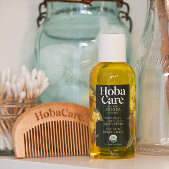 bottle of HobaCare Organic golden jojoba oil surrounded by various bathroom items including a wooden beard comb