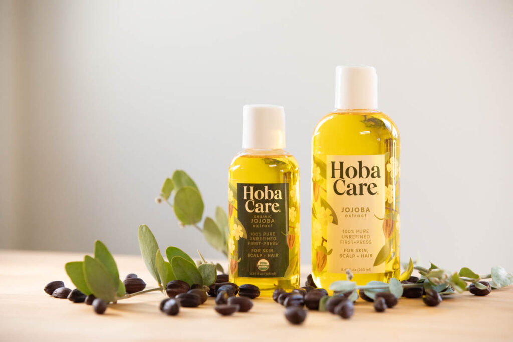 two bottles of golden HobaCare jojoba with jojoba seeds and leaves