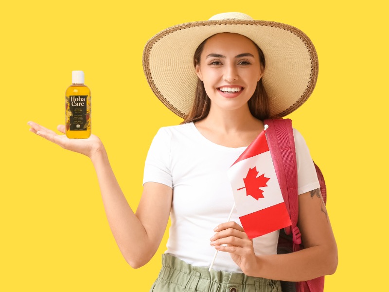 woman in large hat on yellow background holding a Canadian flag in one hand and a bottle of golden HobaCare Jojoba in the other