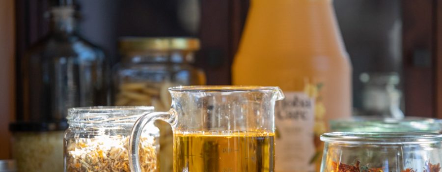 Close-up of jojoba oil in a glass beaker, with dried herbs and flowers in the background.