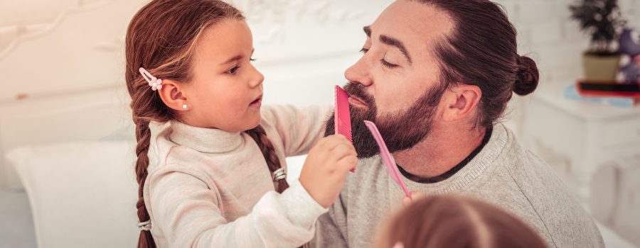 little girl with brown braids combing dad's brown beard with a pink comb to promote that jojoba is the best beard oil